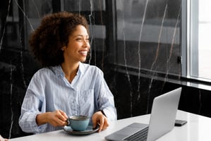 Smiling woman at a laptop with a coffee, looking thoughtfully confident about her career path.