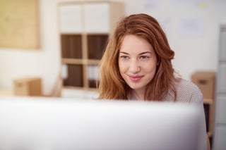 Attractive young woman working on a desktop computer smiling as she leans forwards reading text on the screen, view over the monitor-1