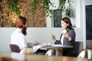 A woman in a grey blazer gestures as she speaks to a bearded man across a small table in a bright café-style office. He listens and takes notes beside coffee cups and an open notebook. Hanging greenery and exposed brick create a relaxed, professional setting suggesting a job interview or coaching talk.