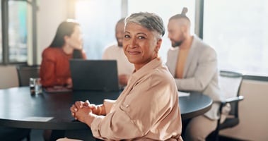 A confident middle-aged woman with short grey hair sits at a round office table, smiling at the camera. Behind her, three colleagues are engaged in conversation with a laptop open in front of them. The scene is brightly lit by natural sunlight streaming through large windows, creating a warm and professional atmosphere that conveys collaboration and confidence in a modern workplace.