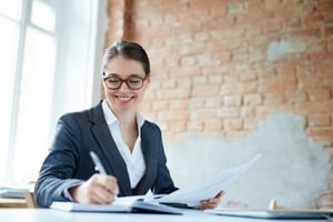 Smiling young woman in business attire writing notes while reviewing documents, representing a career path in accounting.