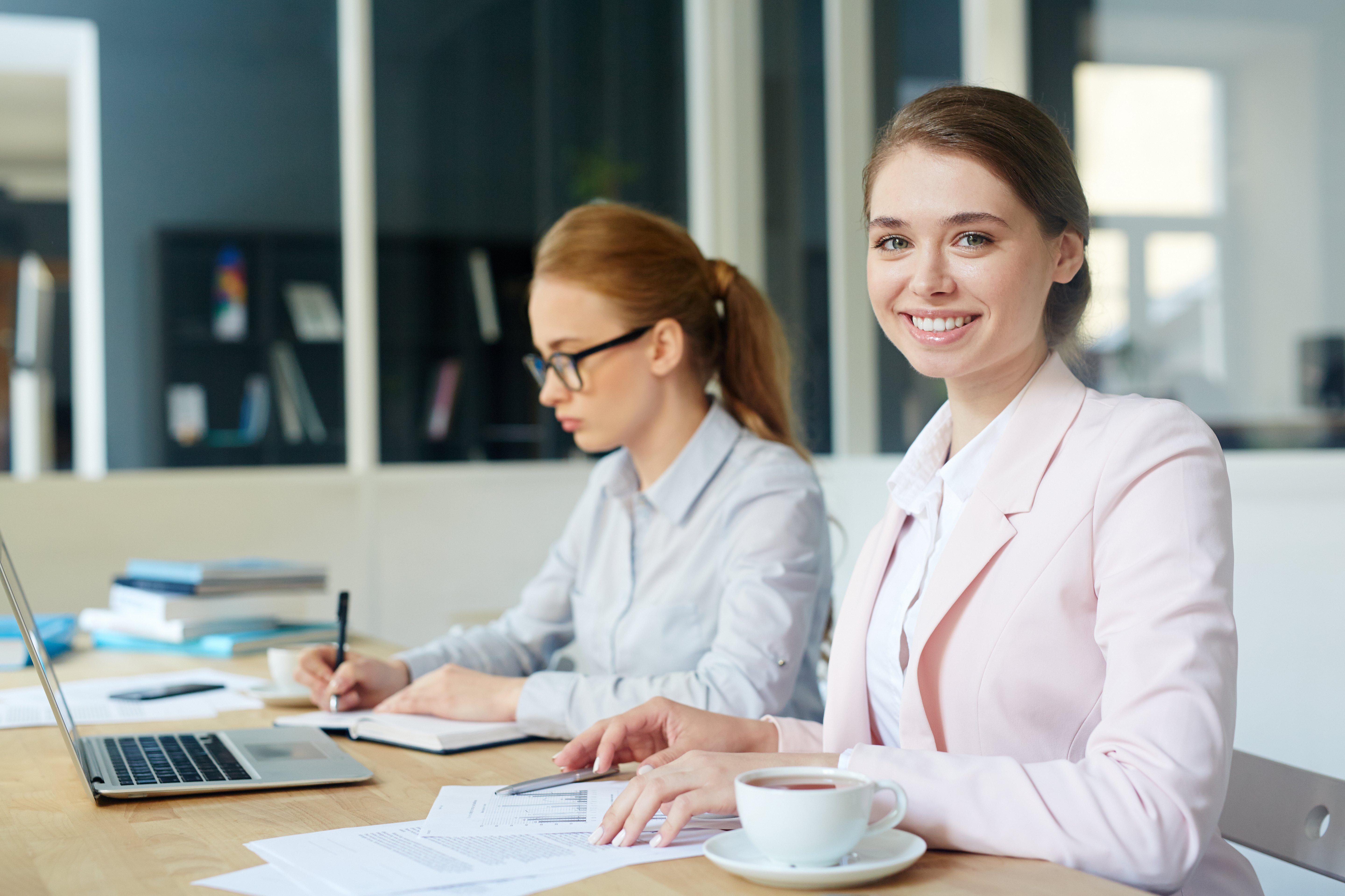 Two young women working at a desk with financial documents and a laptop, representing the learning process of becoming an accountant