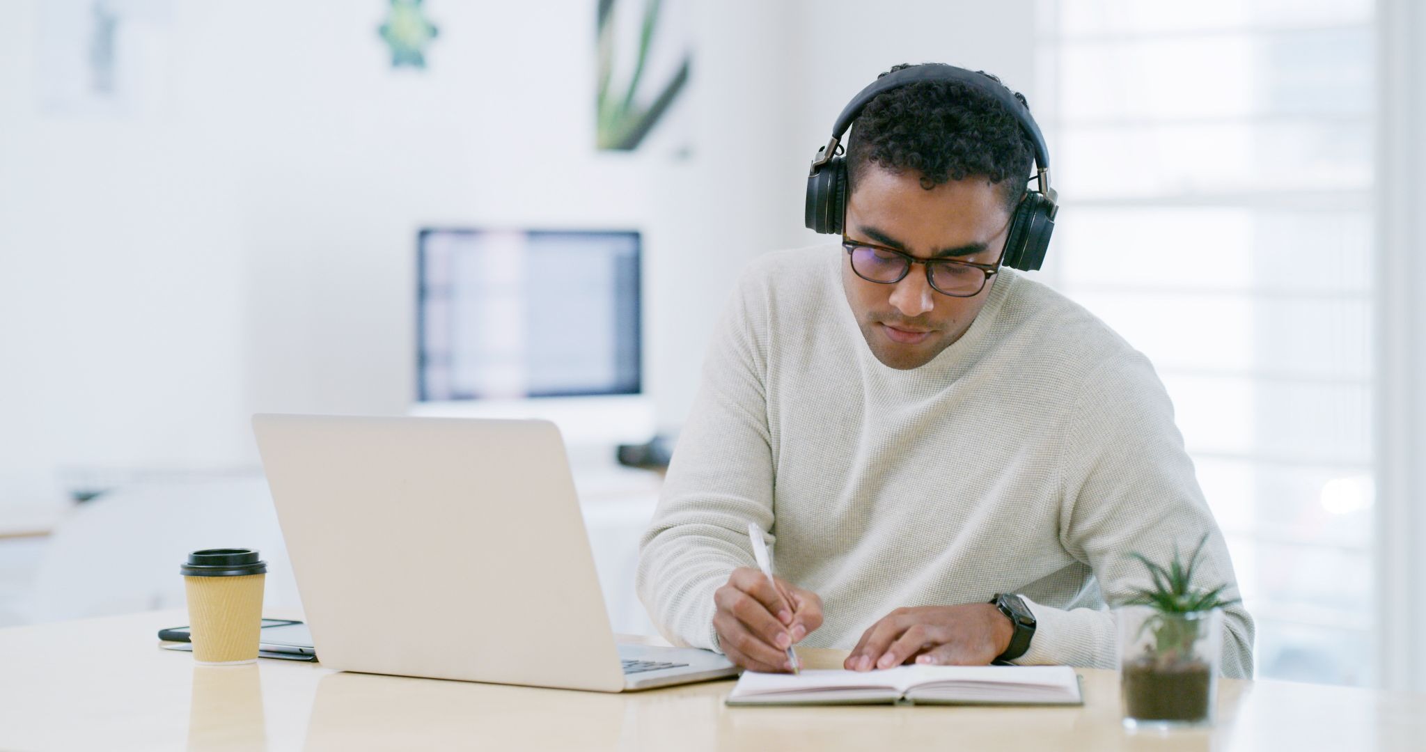 A man wearing headphones studying at a laptop and writing in a notebook, representing flexible online learning.