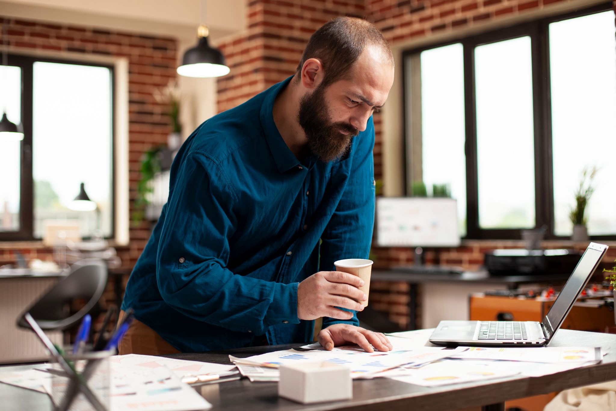 An overworked man leaning over a desk, reviewing paperwork and looking at his laptop while holding a coffee cup.