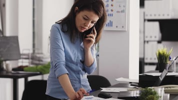 A woman in a blue shirt looking focused and overwhelmed while reviewing documents and taking a phone call at her desk.