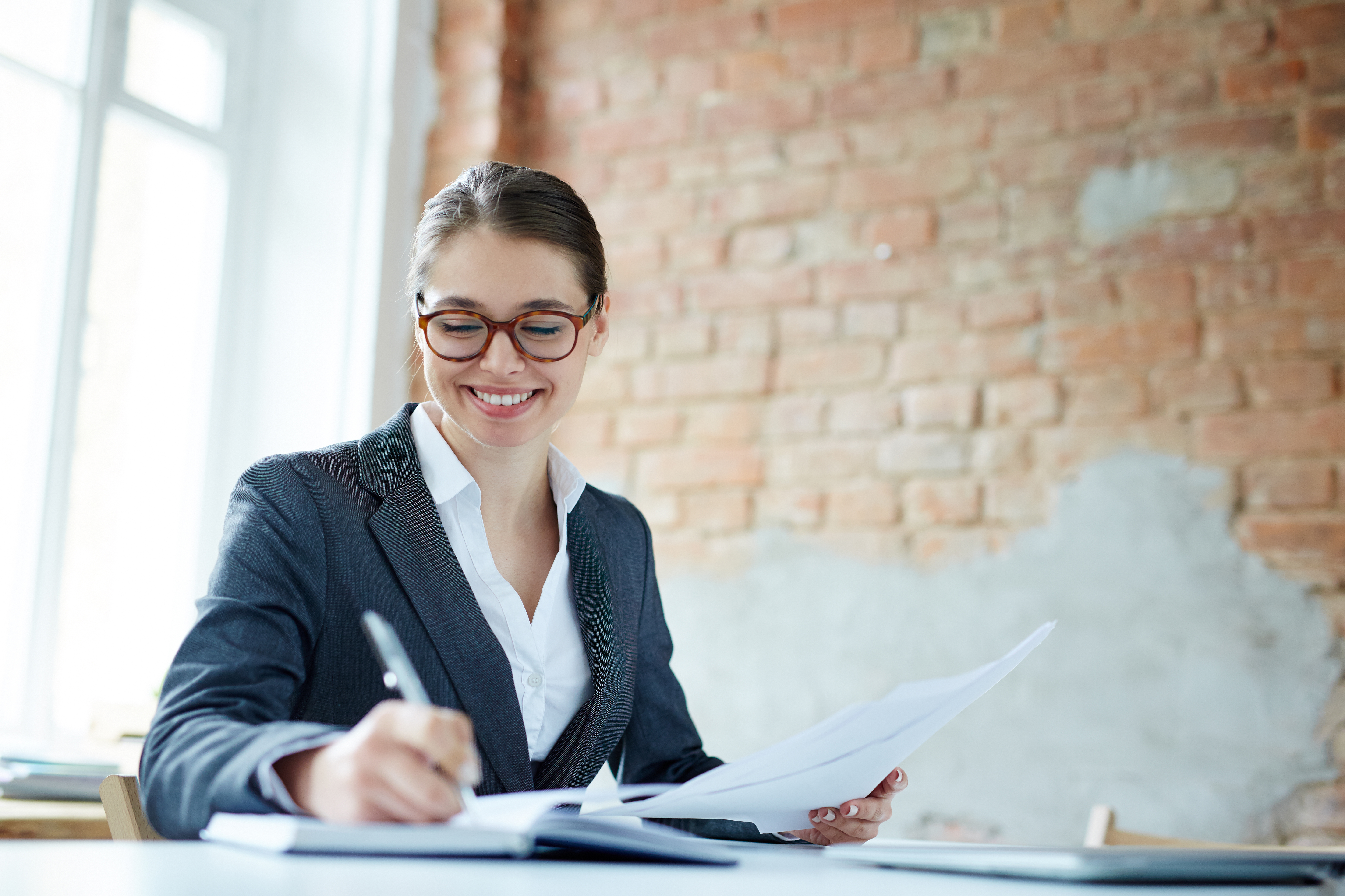 Smiling young woman in business attire writing notes while reviewing documents, representing a career path in accounting.